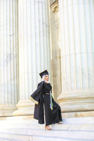 Muslim woman in black graduation gown and cap stands proudly on steps beside tall marble columns.
