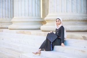 Muslim woman in graduation gown and hijab sitting on marble steps holding cap, celebrating academic achievement.