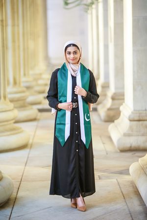 Young Muslim woman in black abaya and hijab wearing Pakistani flag graduation sash, standing between elegant columns.