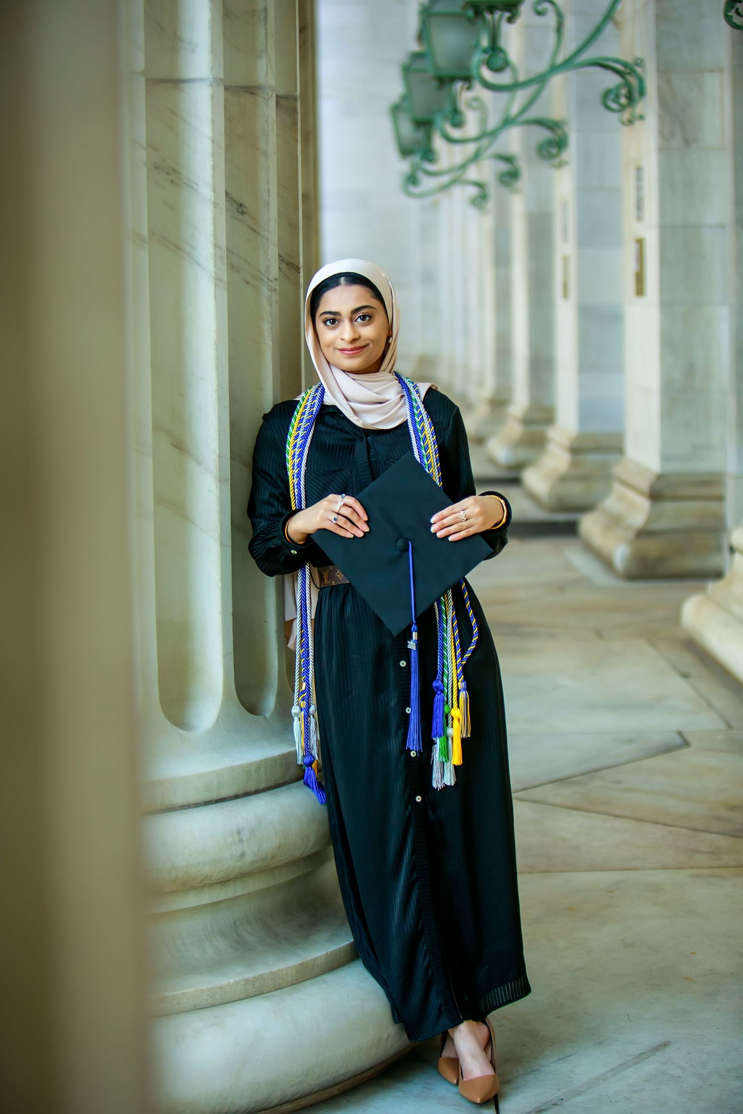 Muslim woman in hijab and black graduation gown holds cap while leaning against columns at university.