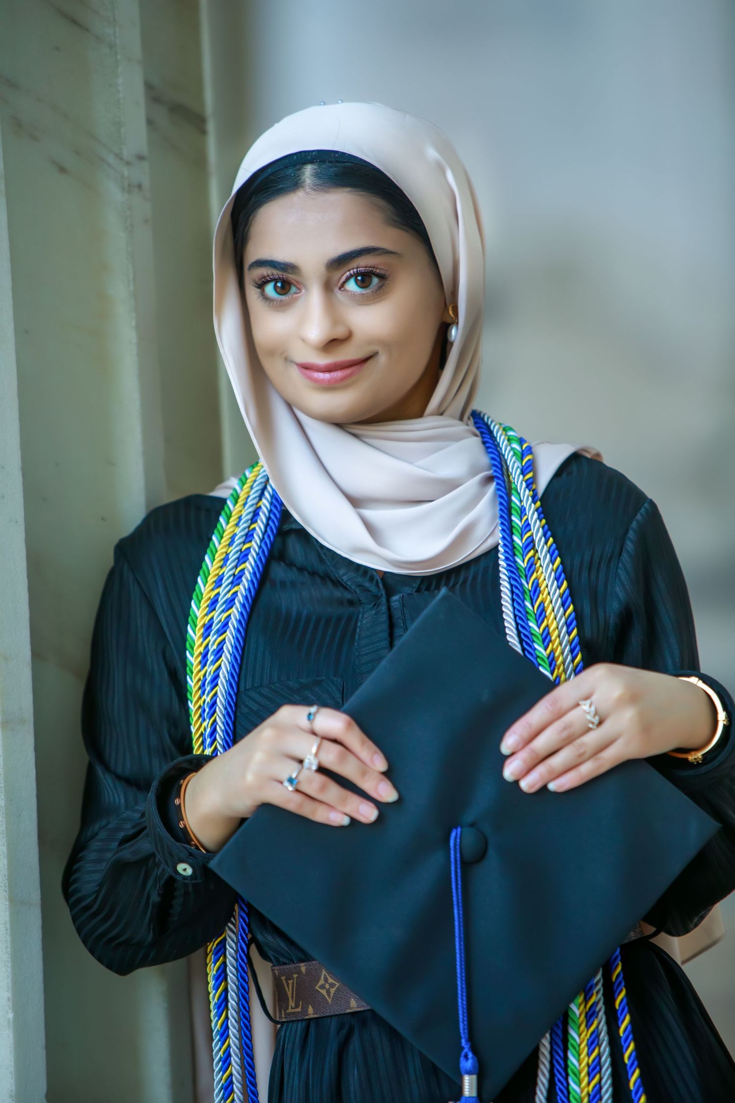 Young woman in hijab and graduation gown holding cap with colorful honor cords, smiling at camera.