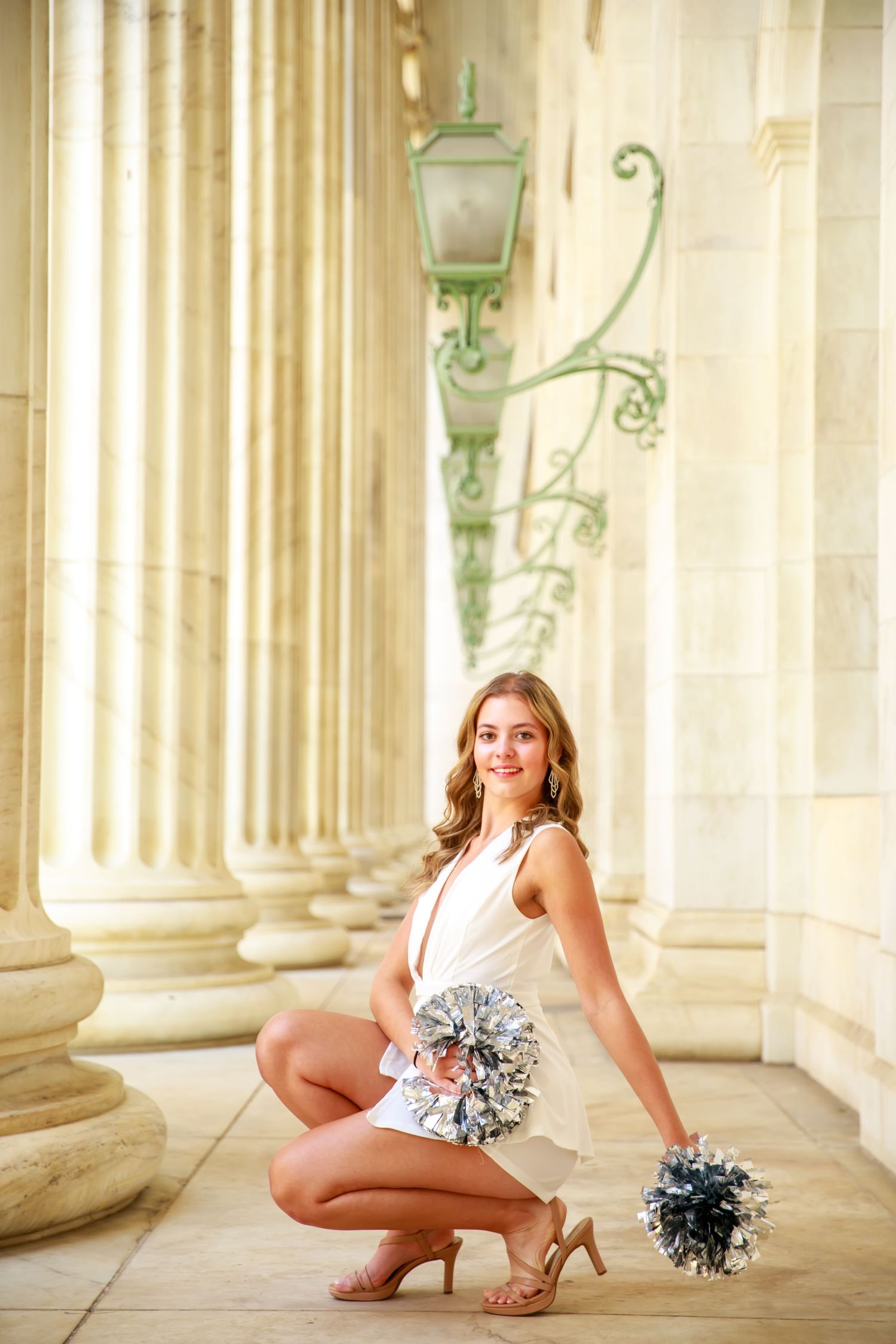 A cheerleader in white uniform kneels with silver pom-poms between ornate stone columns with vintage lanterns.