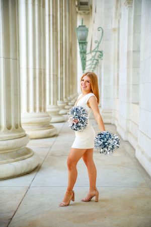 A cheerleader in a white dress holds silver pom-poms while posing between elegant stone columns.