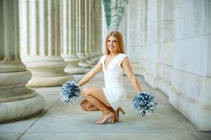 Young cheerleader in white dress poses with silver pom-poms between stone columns.
