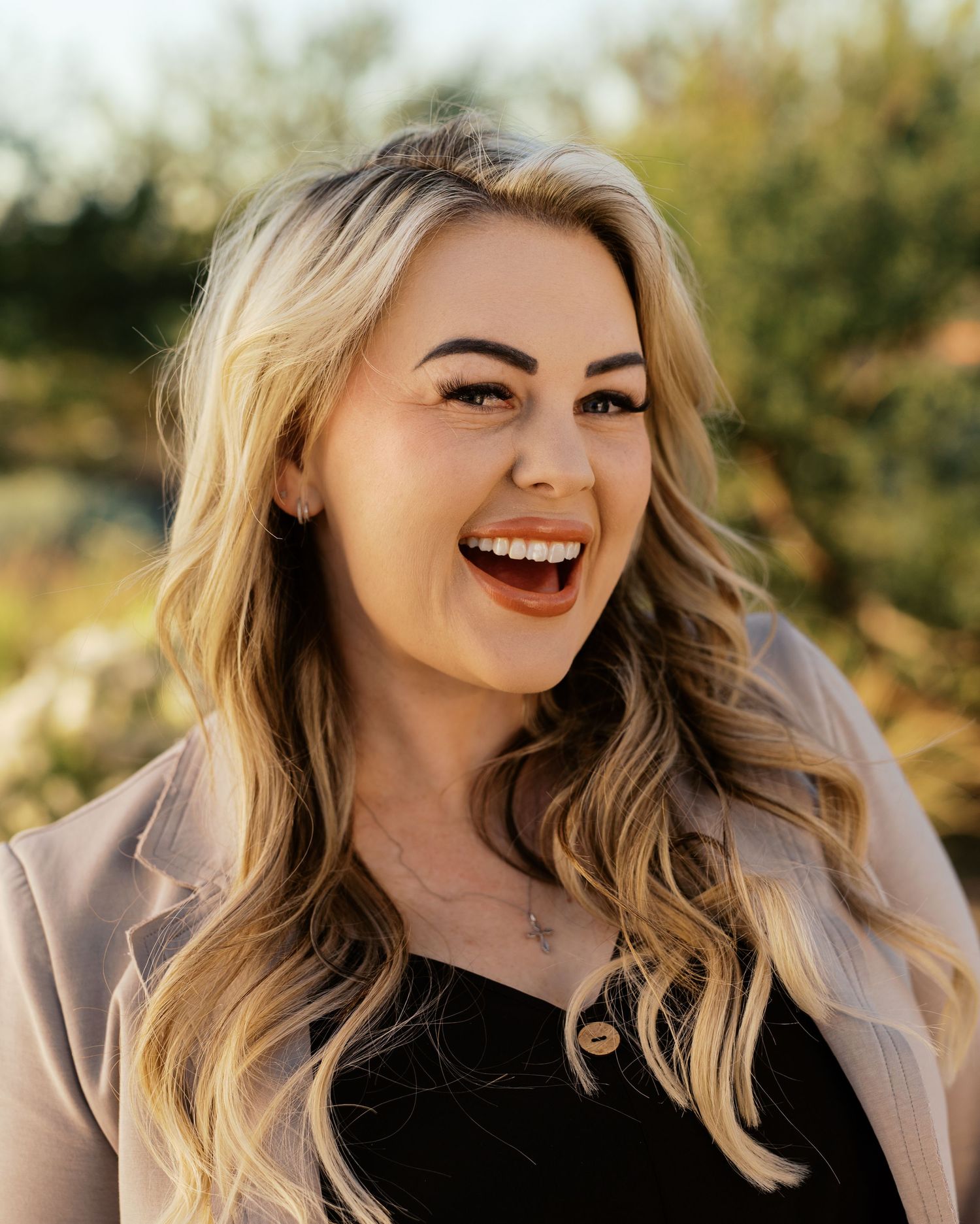 Smiling blonde woman with wavy hair wearing a black top and blazer outdoors in warm golden light.
