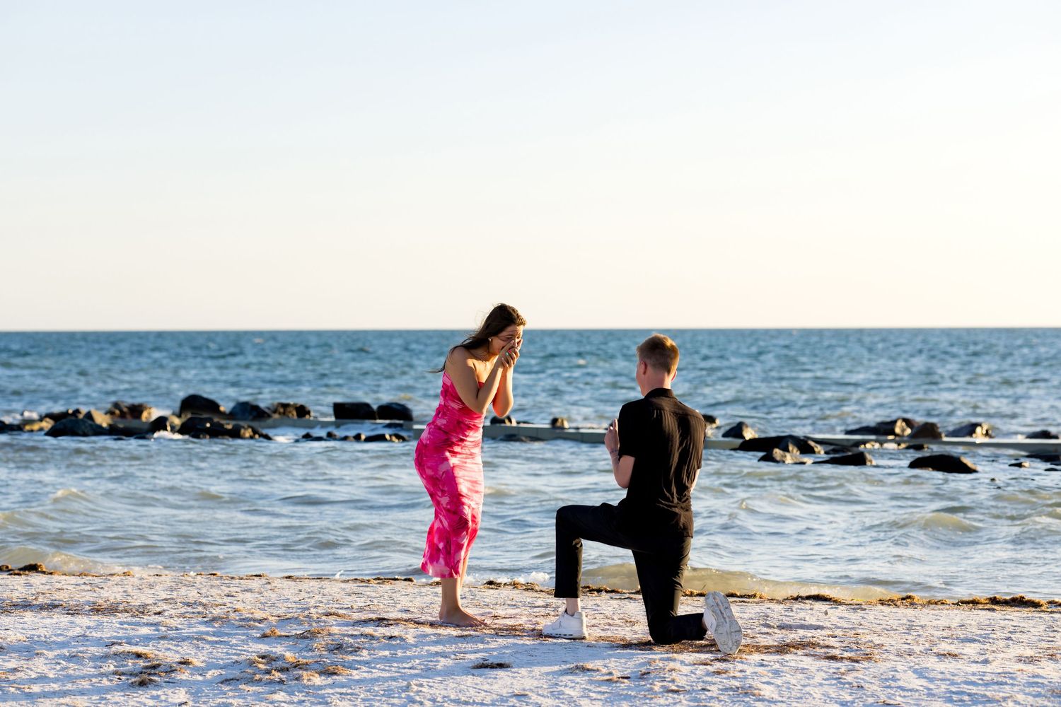 Man proposes on bended knee to woman on a sandy beach at sunset with ocean waves in the background.