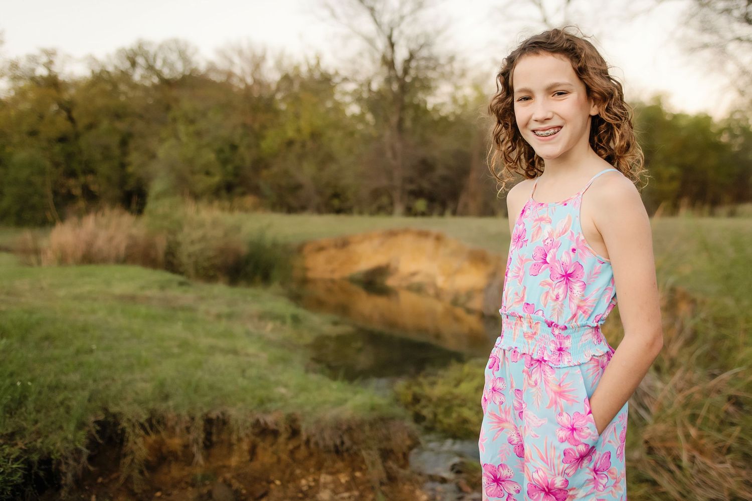 A smiling girl in a floral blue and pink jumpsuit poses near the creek surrounded by lush greenery at The Keller Pointe in Keller, TX.
