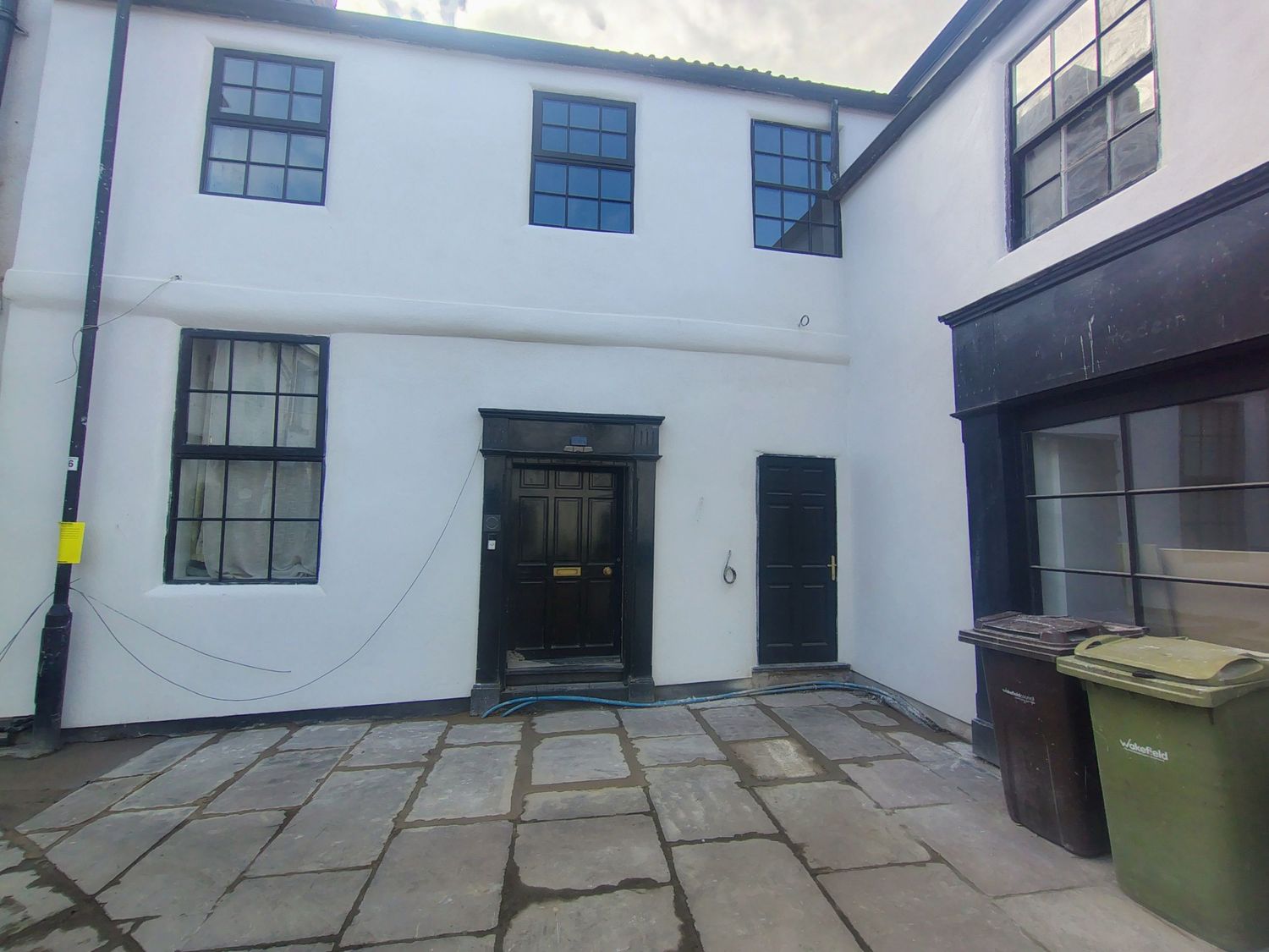 White two-storey building with black-framed windows, dark entrance door, stone paved courtyard, and green wheelie bin outside.