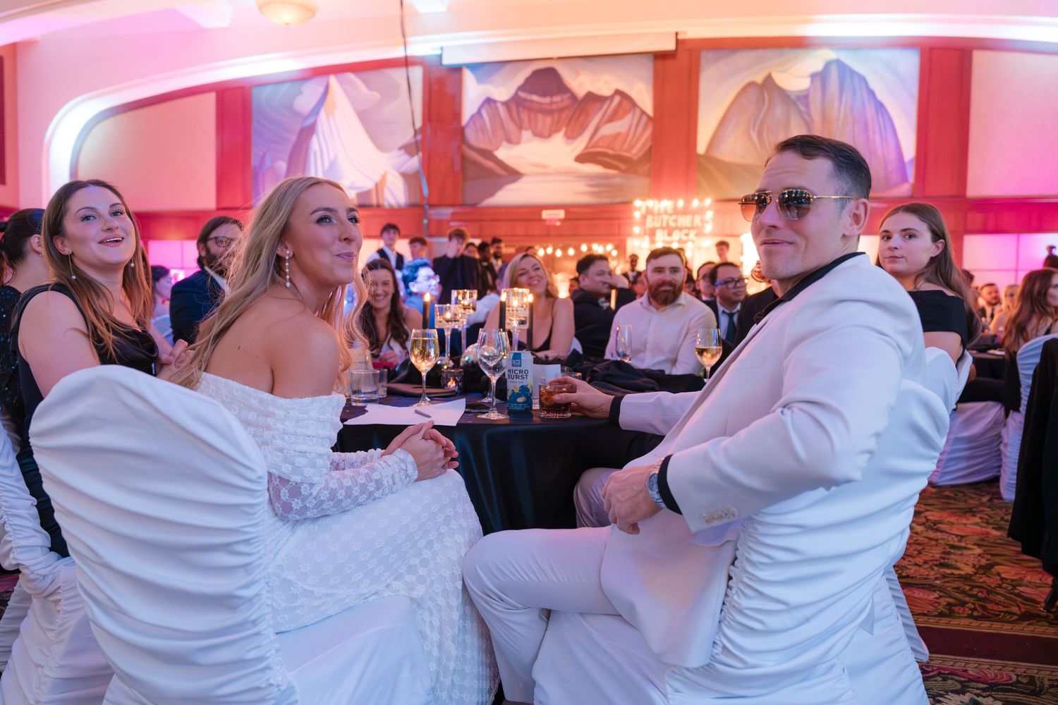 Couple in white attire seated at a gala event table, surrounded by guests in an elegant ballroom.