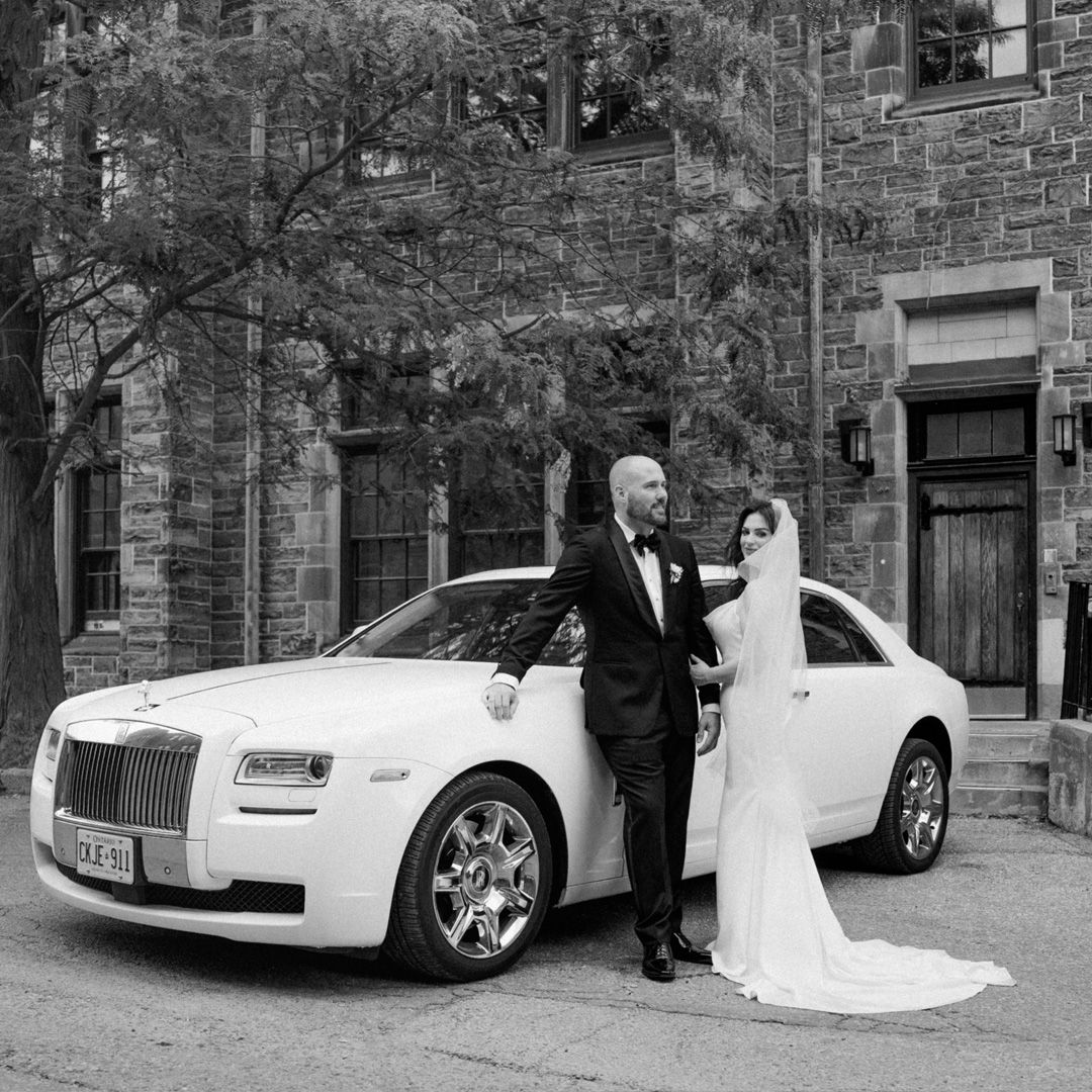 Black and white photo of a bride and groom posing beside a white Rolls-Royce Ghost on their wedding day.