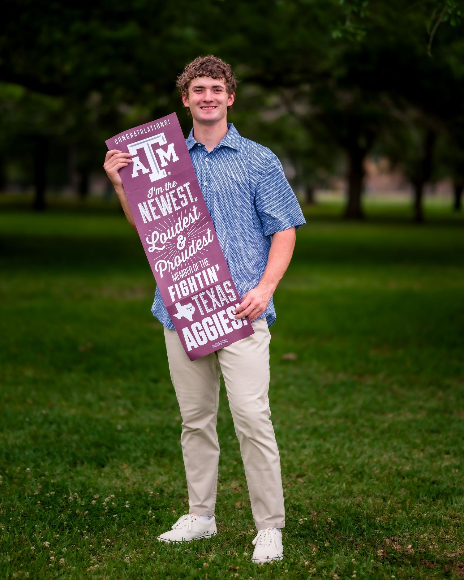 Young man smiling outdoors holding a Texas A&M Aggies banner, wearing a blue shirt and khaki pants on green grass.