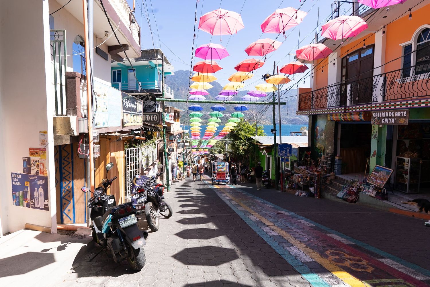 Colorful pink umbrellas suspended above a narrow street create shade and whimsy in a Mediterranean town.