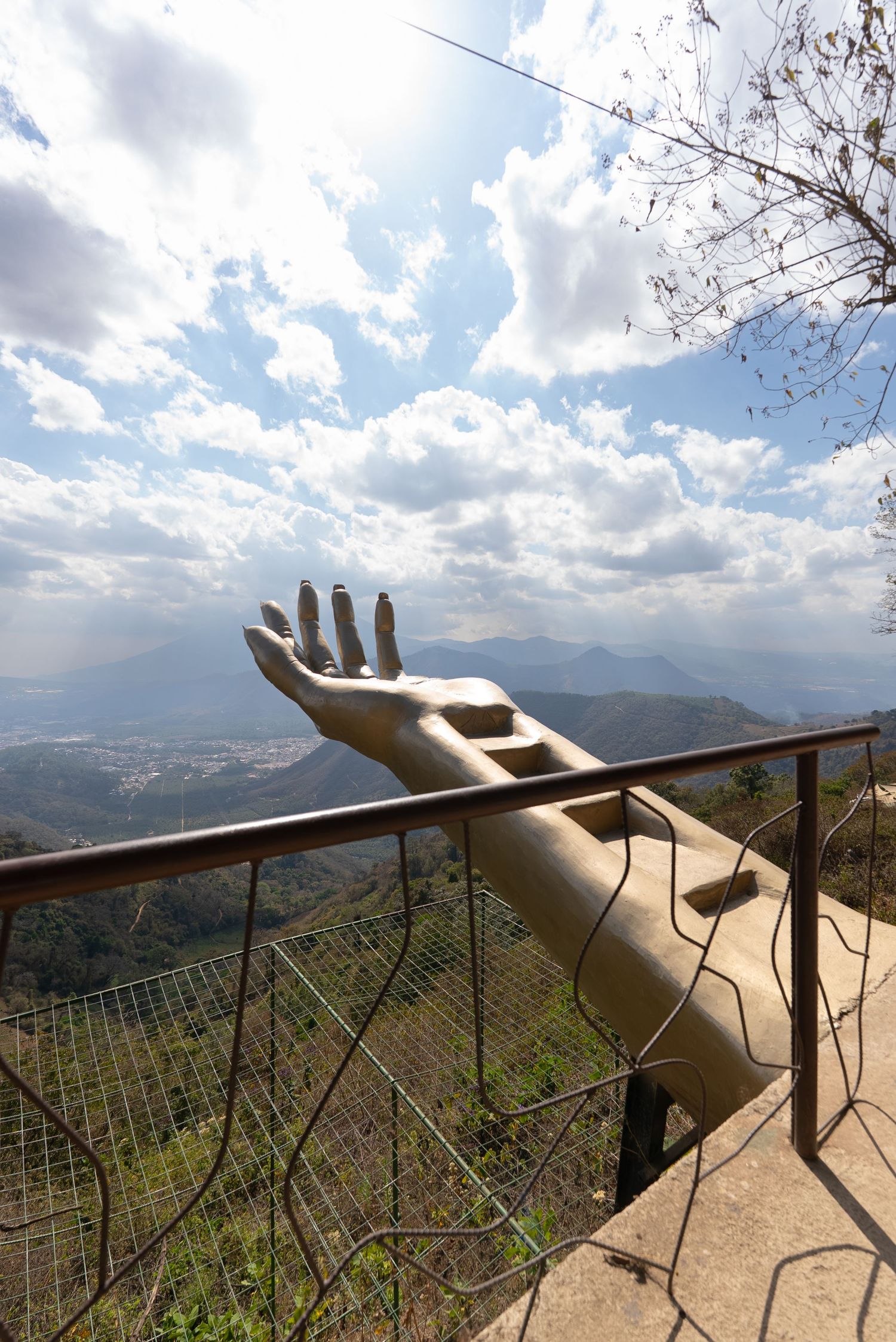 A series of artistic metal hand sculptures reaching outward along a mountain viewpoint railing against a scenic valley backdrop.