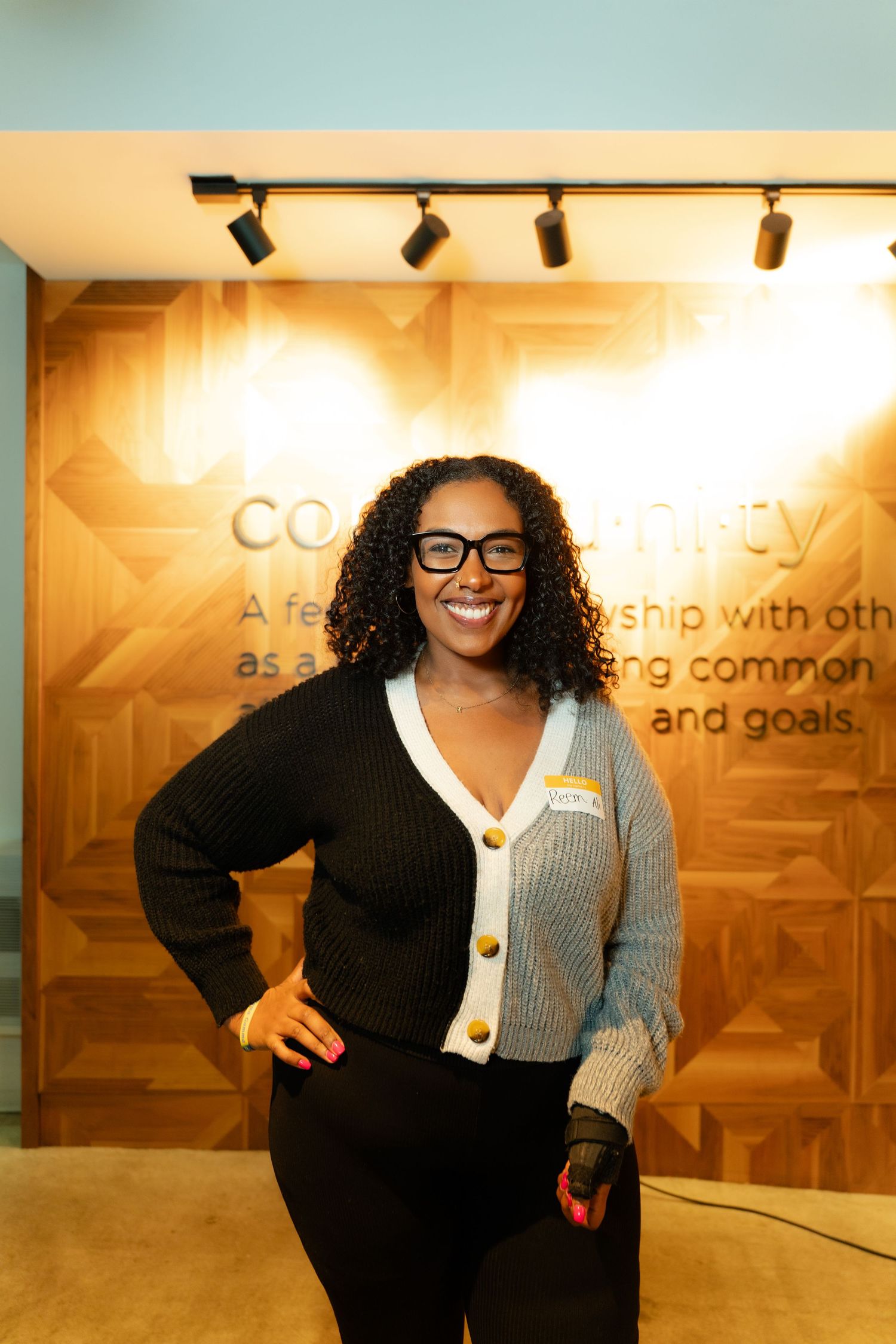 A person in a black blazer and gray sweater poses energetically in front of a corporate office wall display.