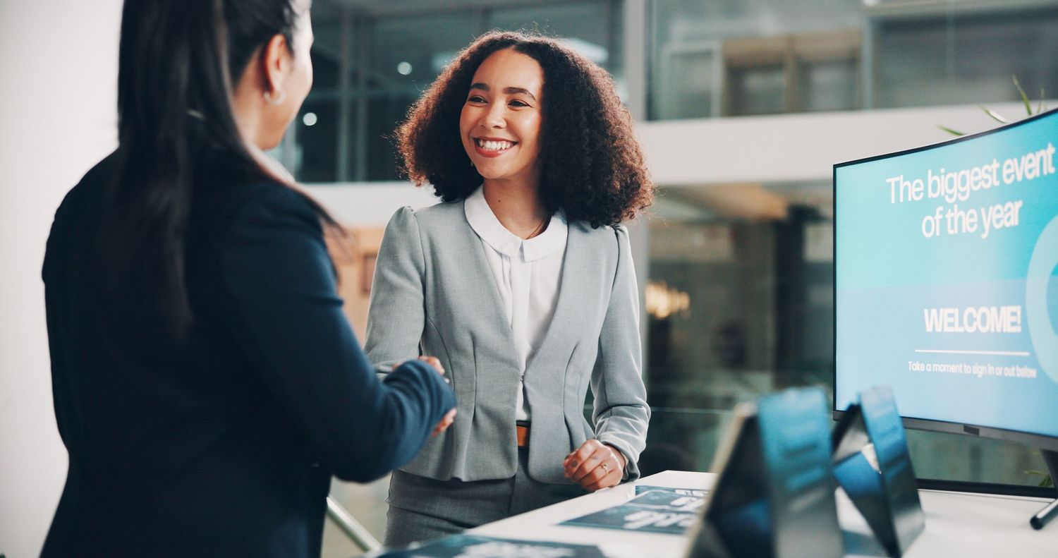 Two businesswomen shaking hands at a reception desk with a welcome screen in the background.