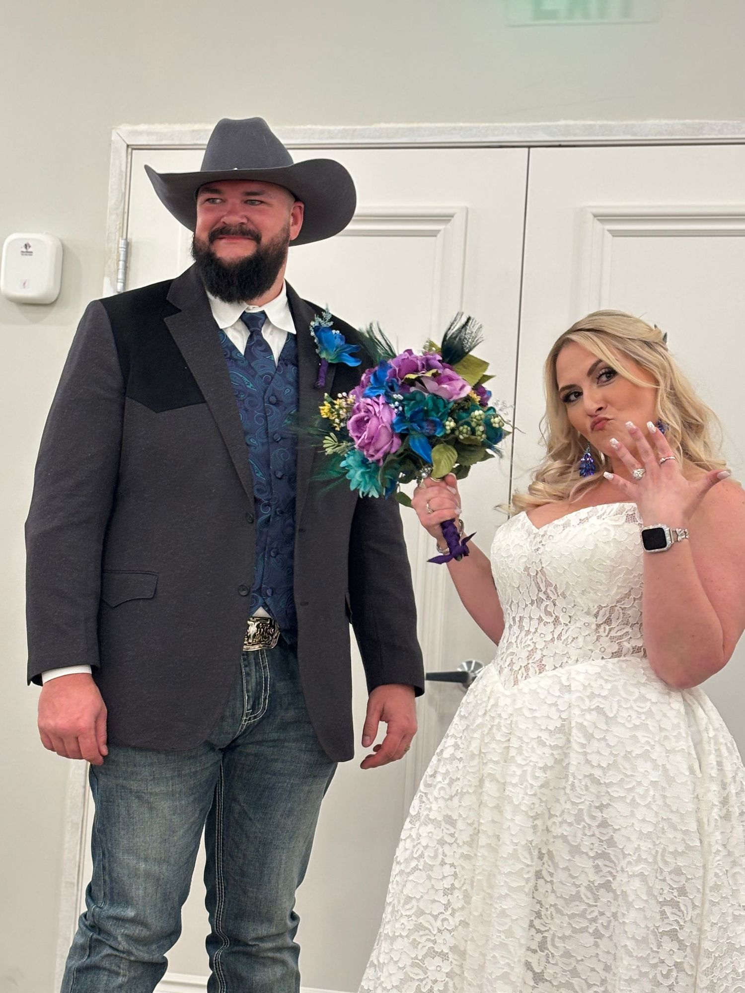 Newlywed couple posing together, groom in cowboy hat and suit, bride in white lace gown holding colorful bouquet.
