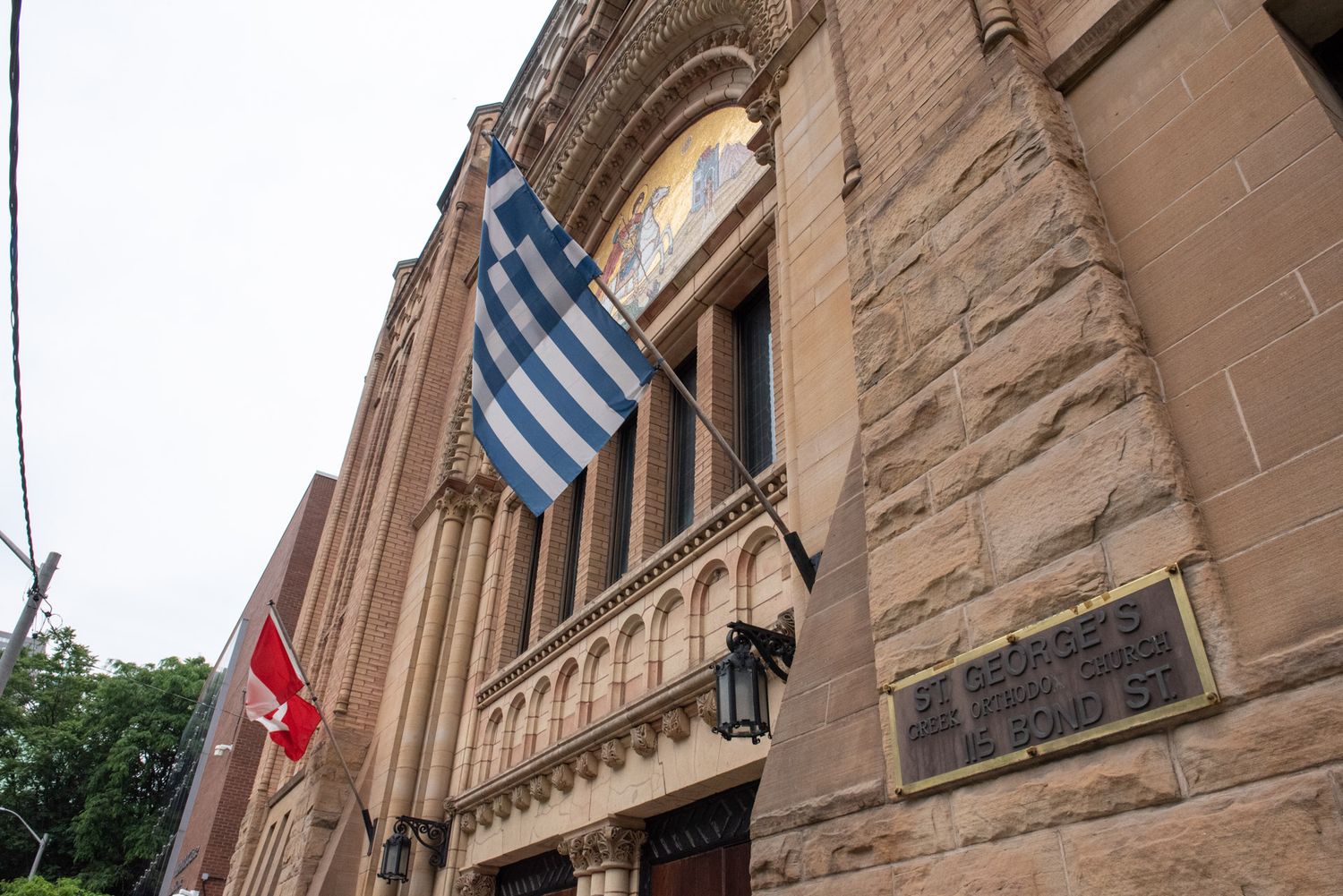 A Greek Orthodox church exterior with a blue and white Greek flag displayed on the stone building facade.