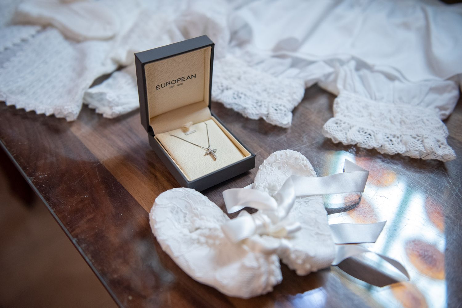 A gold cross necklace in a jewelry box surrounded by white lace baby booties and christening garments.