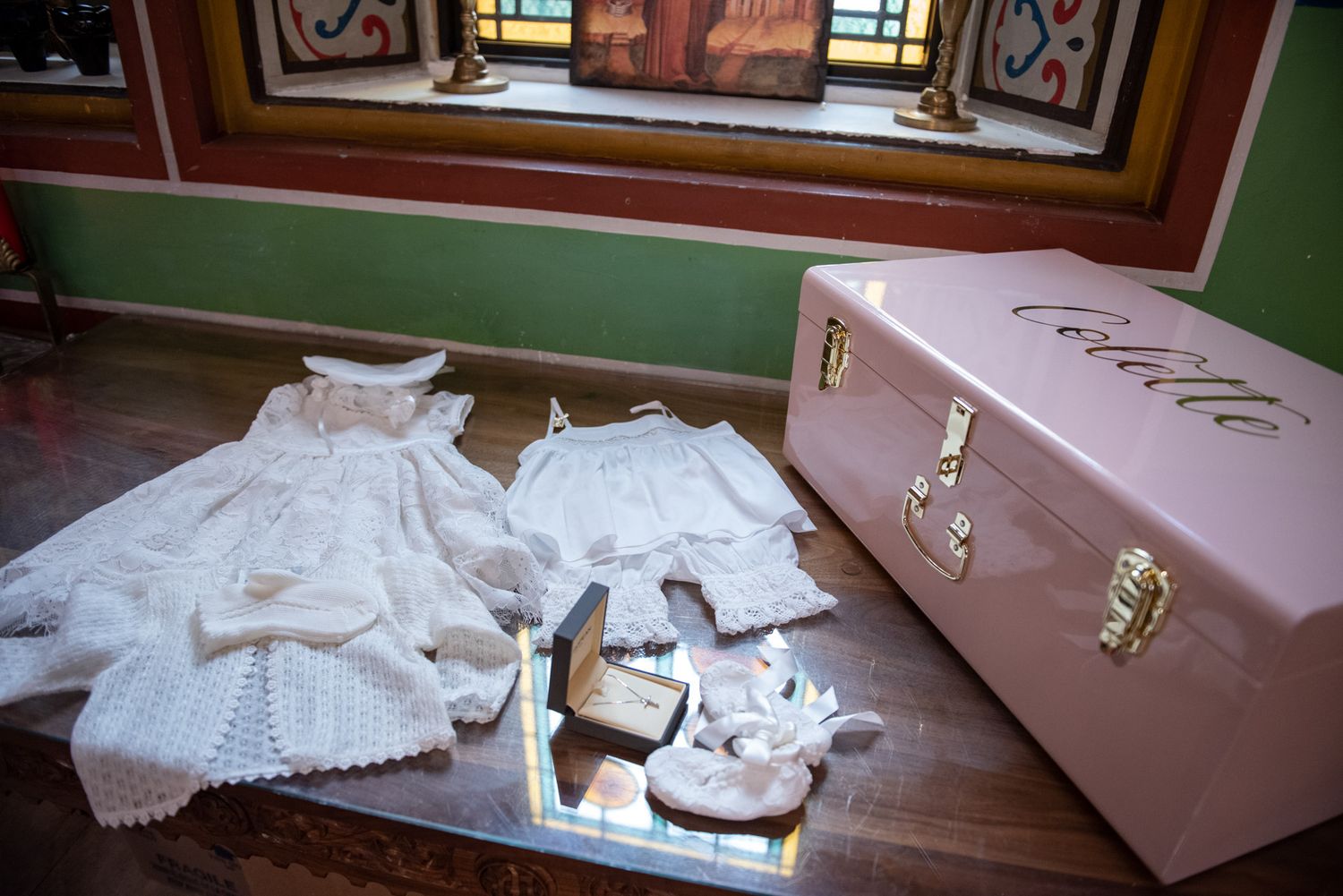 White lace christening gown and accessories laid out beside a pink personalized Colette keepsake trunk.