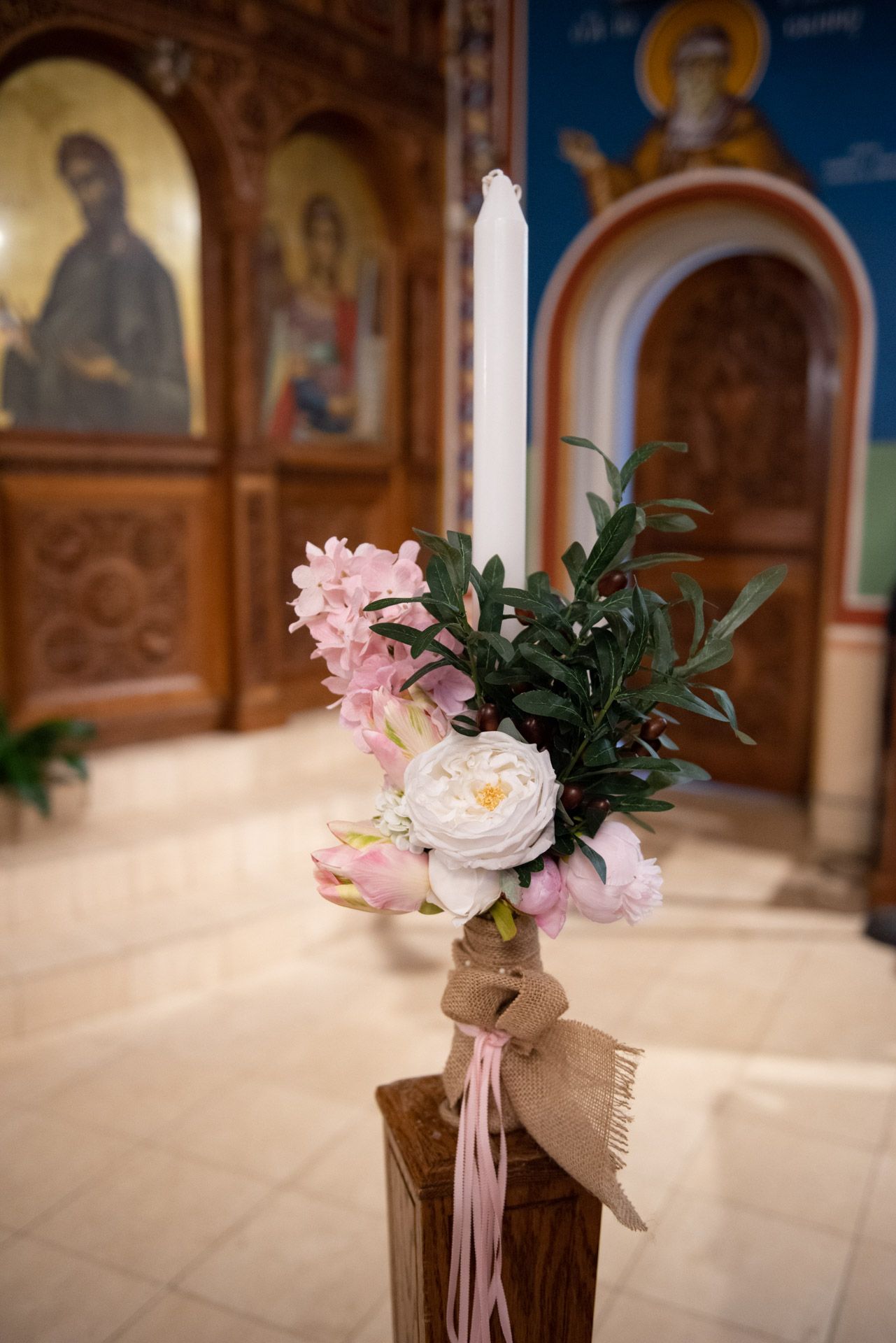 An elegant white baptism candle adorned with pink and white flowers tied with a burlap ribbon in a church.