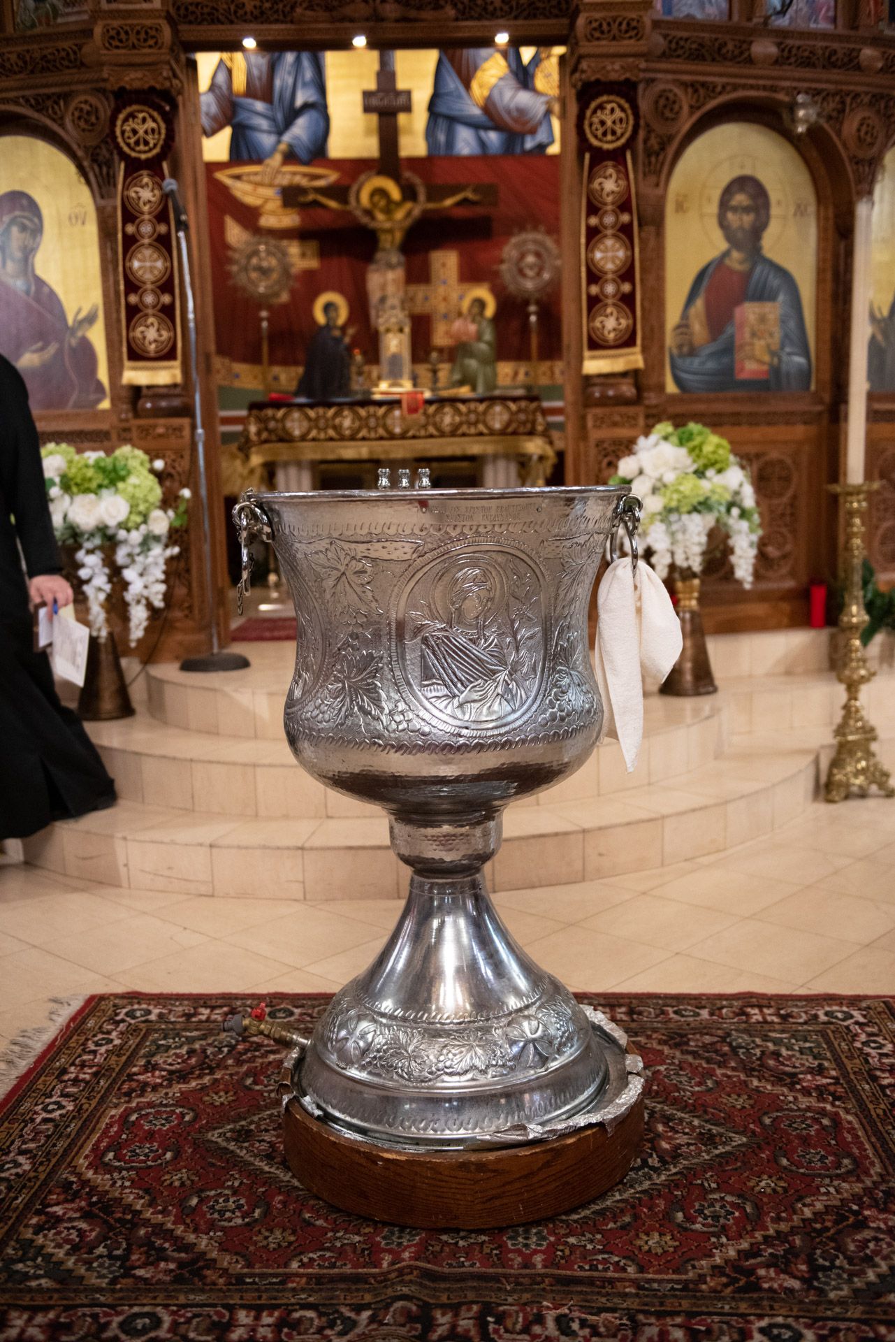 An ornate silver Greek Orthodox baptismal font standing on a wooden base inside a decorated church.