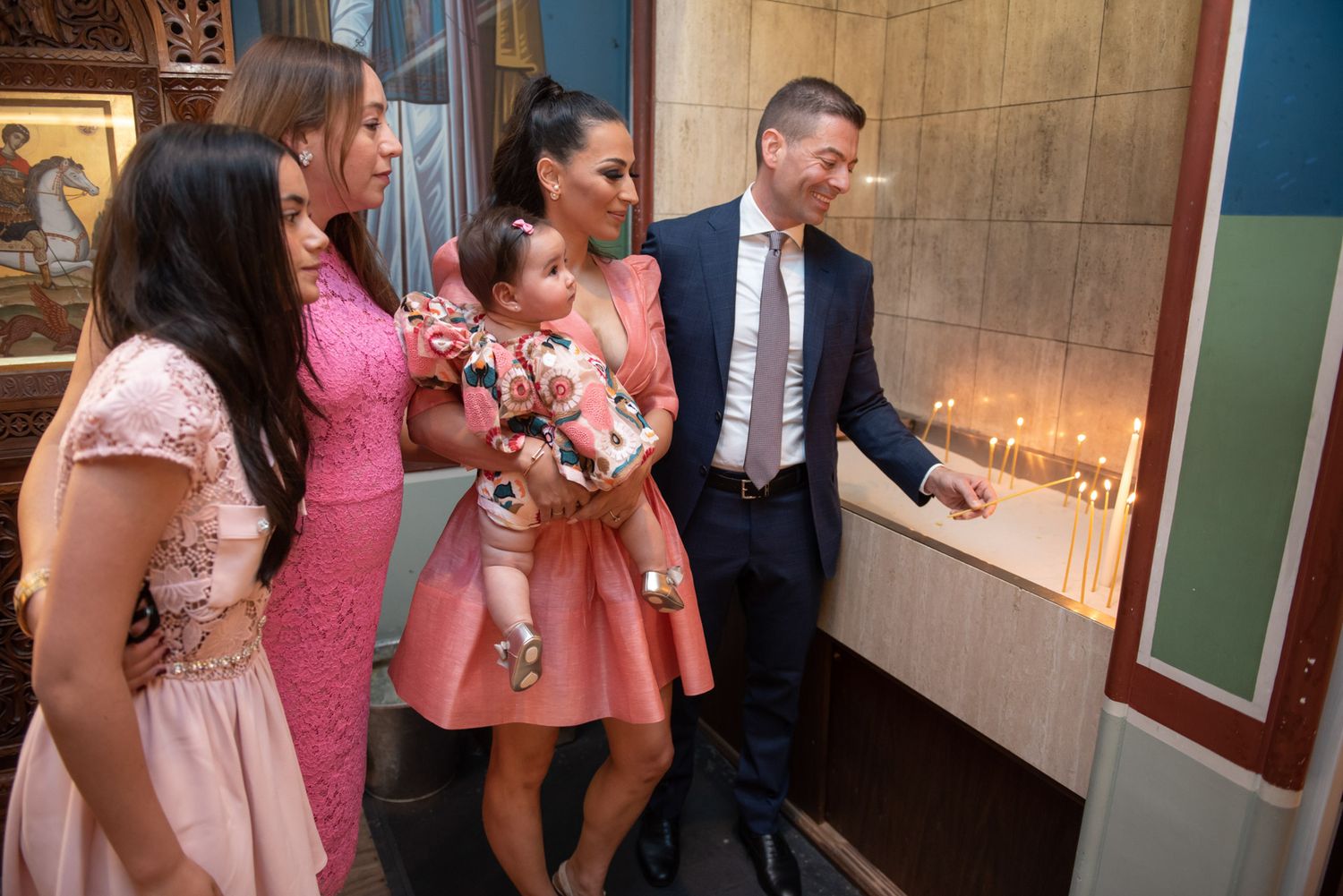 A family lighting candles at a Greek Orthodox church during a baptism ceremony with a baby in pink.