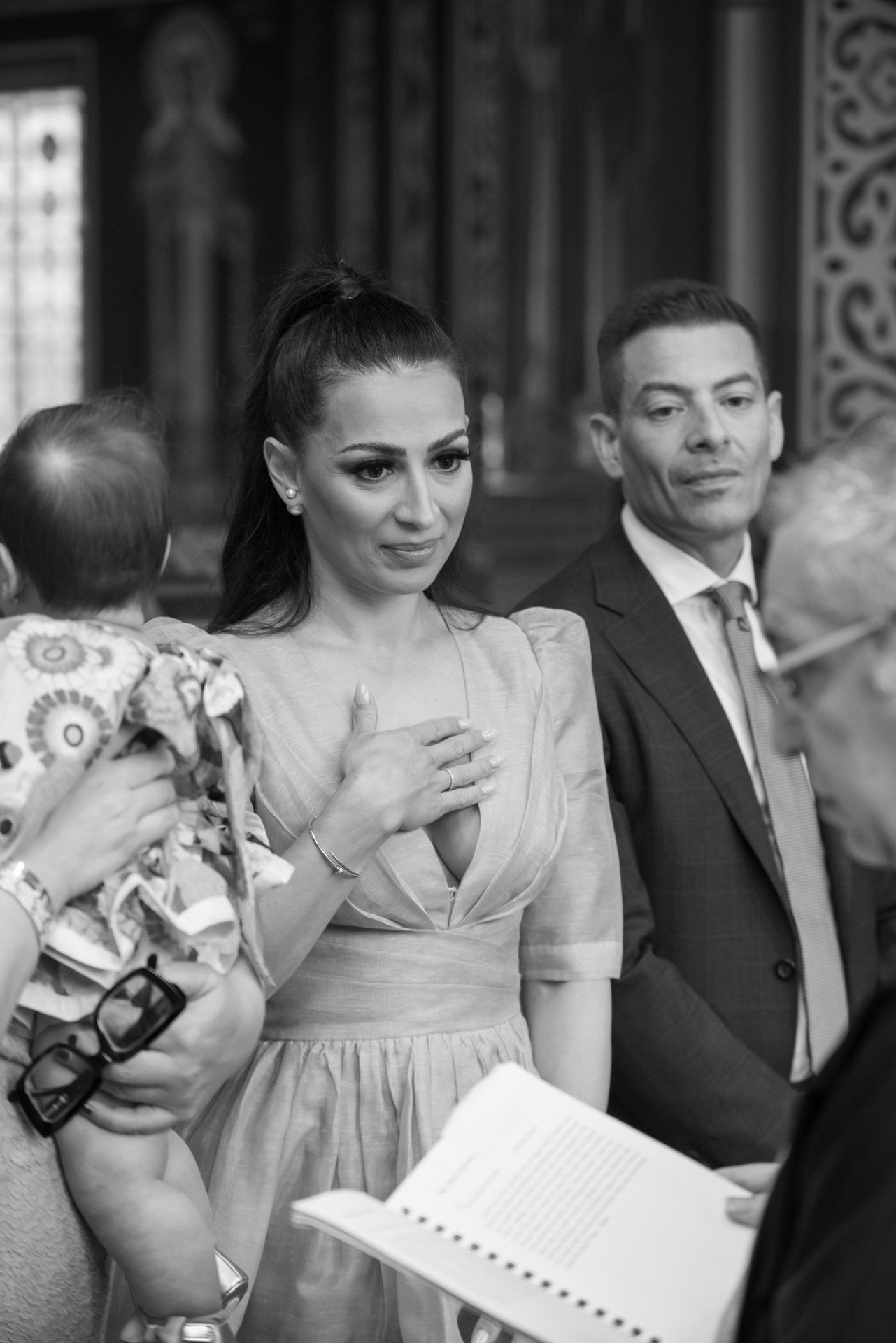 An emotional mother in a beige dress holds back tears during her baby's baptism ceremony in a church.