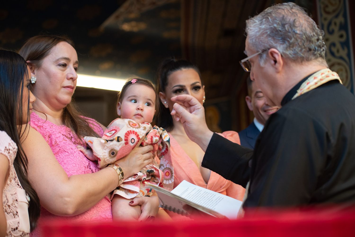Priest anoints baby during Orthodox Christian baptism ceremony while mother holds infant in church.