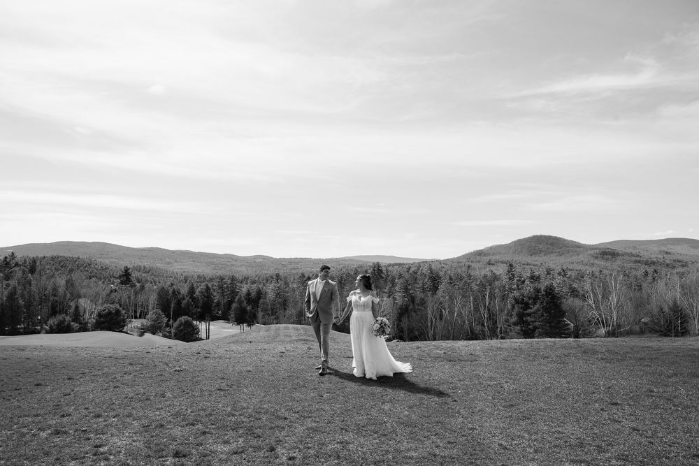 Black and white photo of a bride and groom holding hands walking through a scenic mountain meadow landscape.