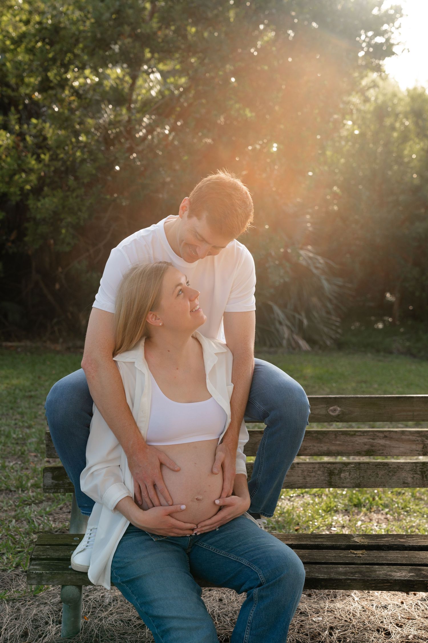 Pregnant couple sitting on park bench in golden sunlight, man embracing woman from behind, both cradling baby bump.