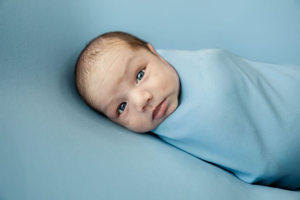 Newborn baby boy swaddled in light blue wrap lying on matching blue backdrop.