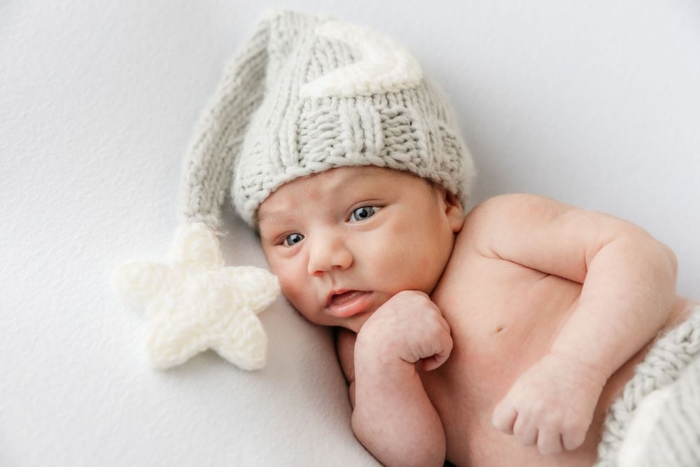 Newborn baby wearing oversized gray knit nightcap with star detail lying on white backdrop.