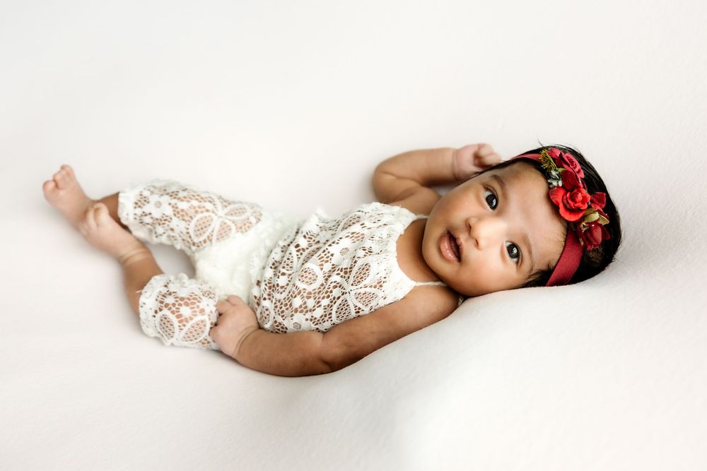 Adorable baby girl in white lace romper and red floral headband, smiling while lying on a white background.