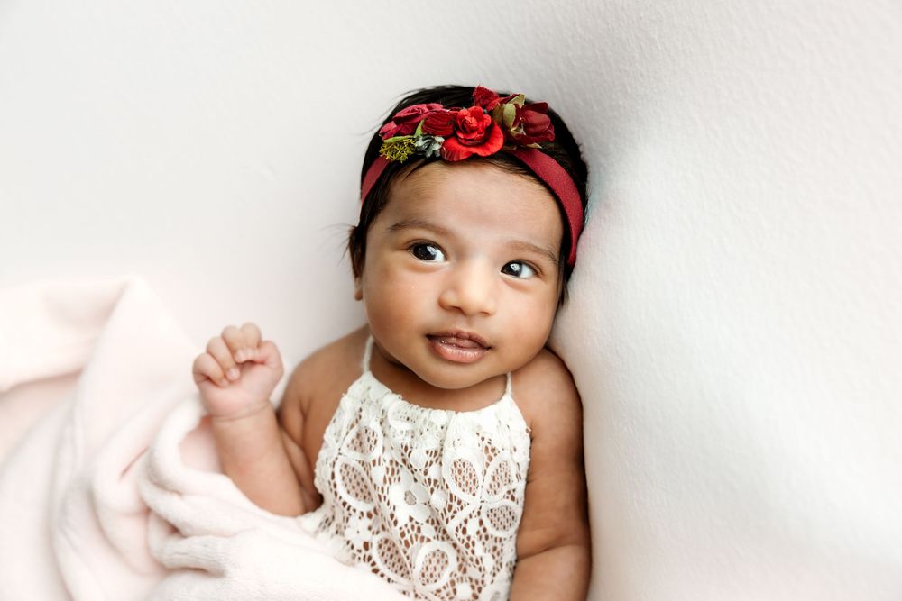 Newborn baby girl in white lace romper and floral red headband lying on soft pink backdrop.