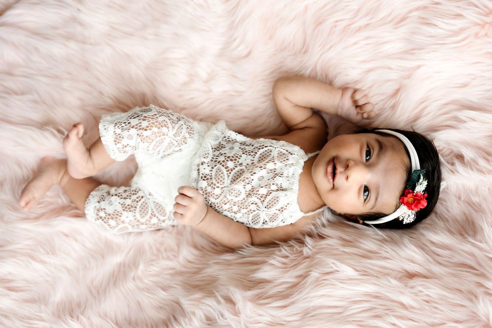Adorable baby girl in white lace romper with floral headband lying on fluffy pink fur rug.