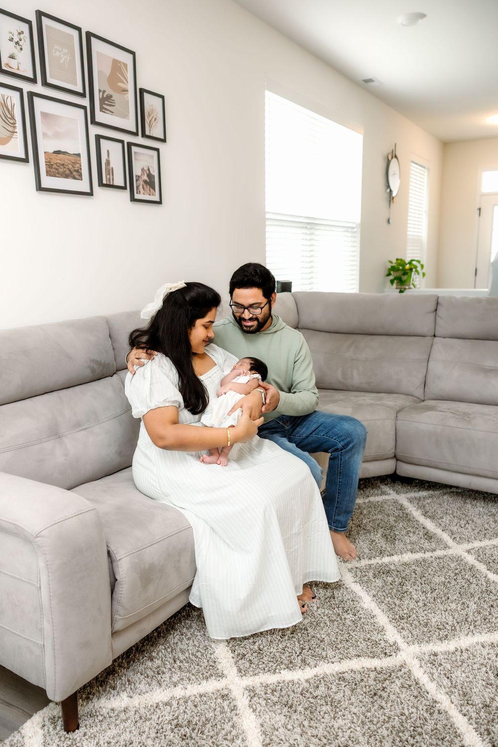 New family of three sitting on gray sectional sofa admiring their sleeping newborn baby.