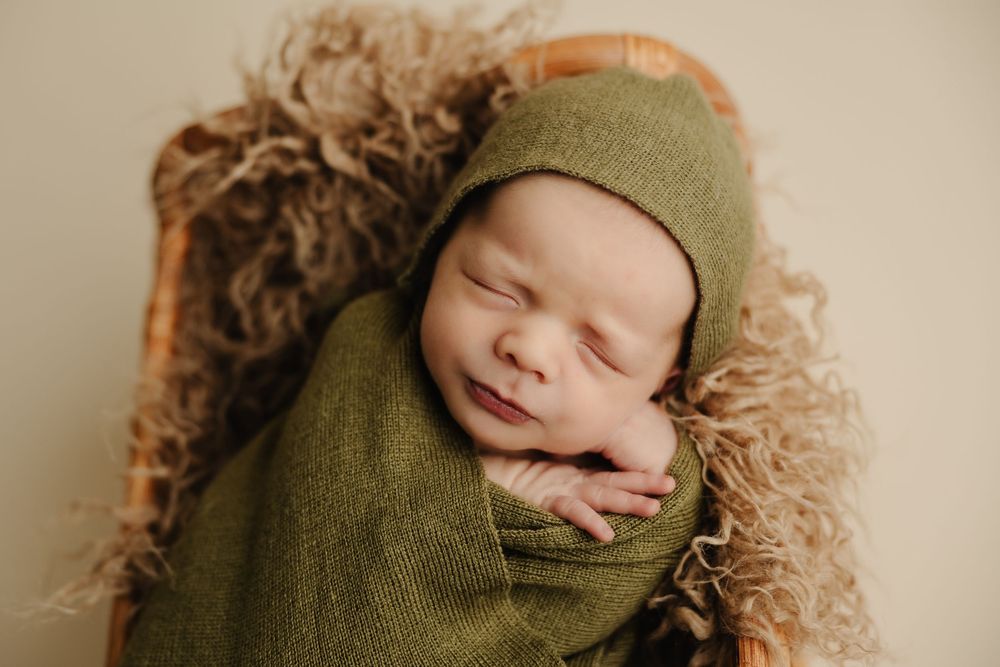 Sleeping newborn baby swaddled in olive green wrap with matching bonnet, resting in a wicker basket with fluffy lining.