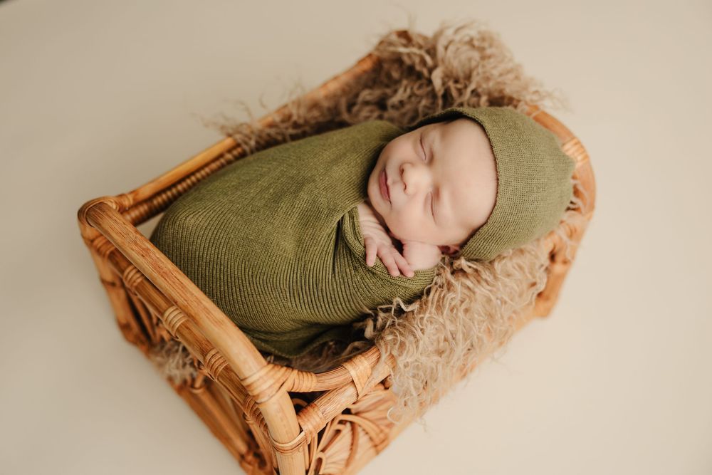 Smiling newborn baby wrapped in green knit swaddle and bonnet, peacefully sleeping in a small wicker basket.