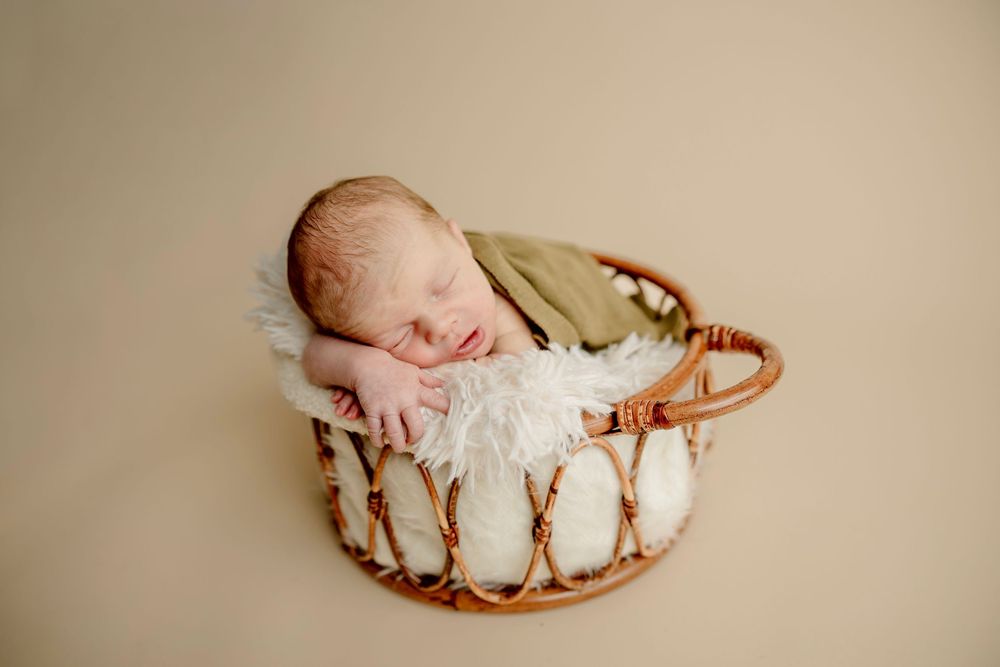 Newborn baby sleeping in a round wicker basket with white fluffy lining, wrapped in a soft olive green blanket.