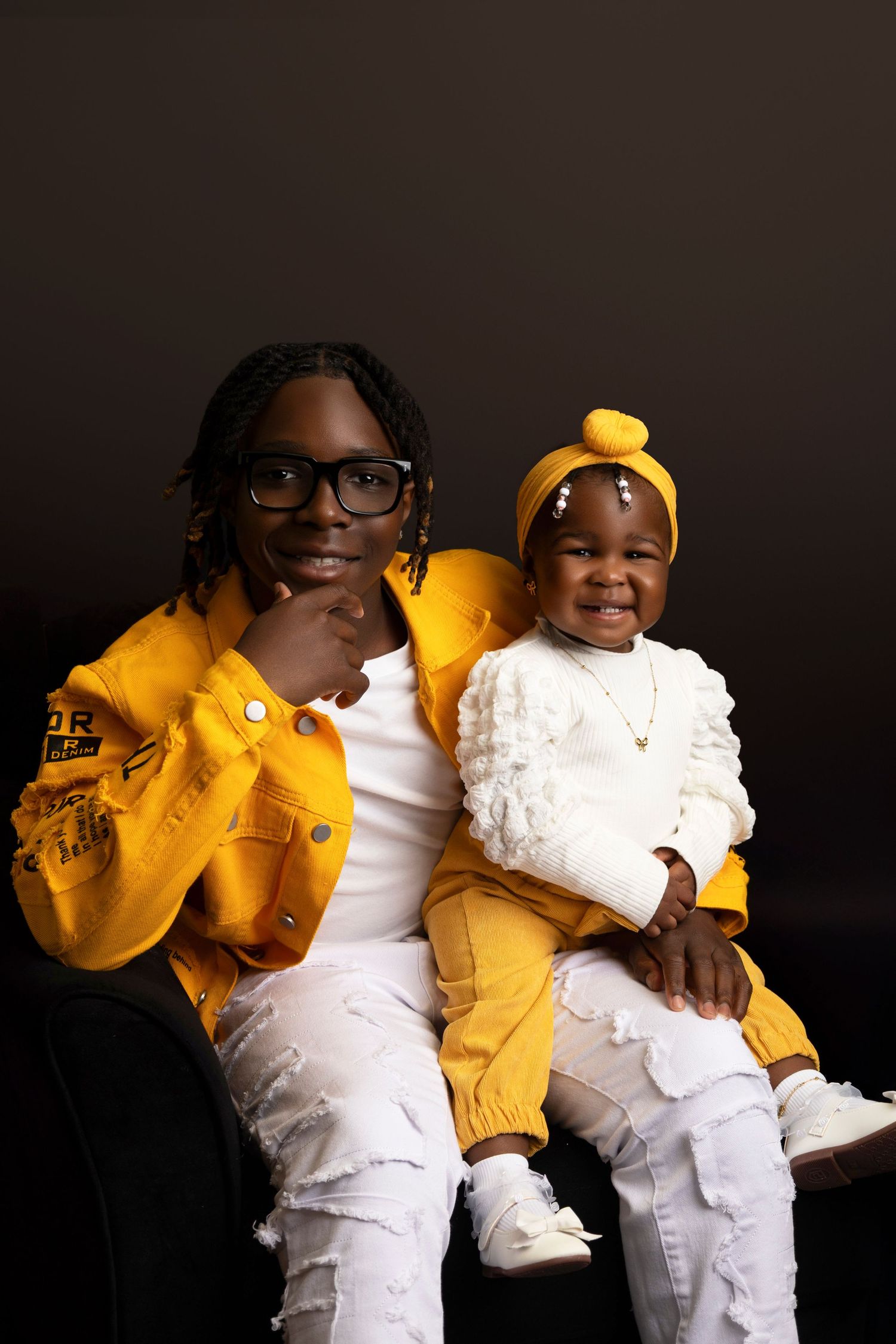 A young man in yellow jacket and white jeans poses with a smiling baby girl in white and yellow outfit.