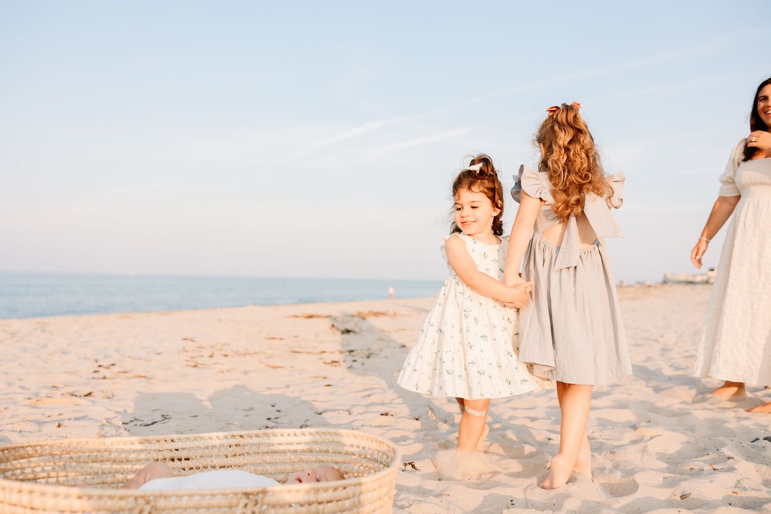 Two young girls in dresses playing on a sandy beach with a wicker basket, ocean in the background.