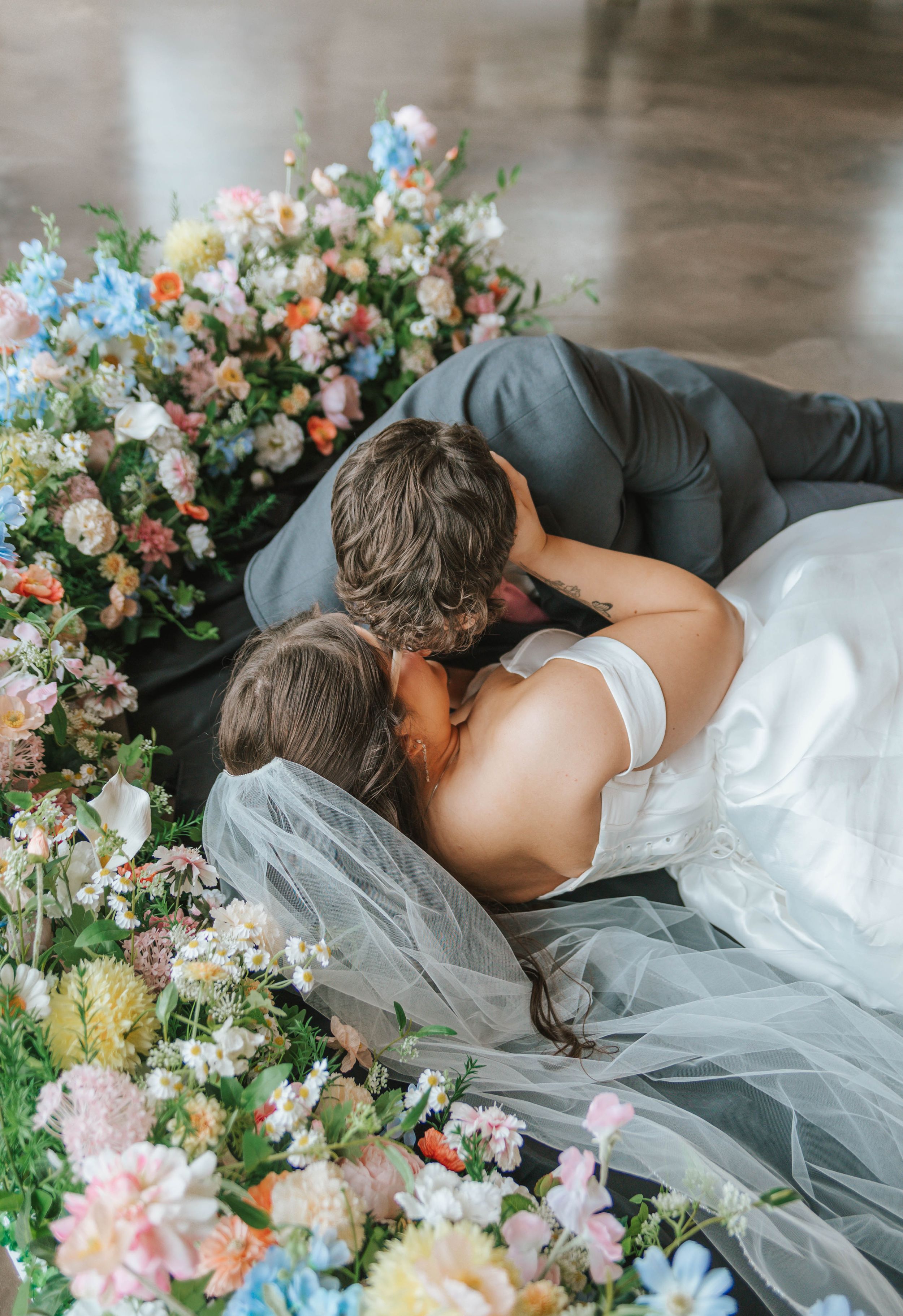Bride and groom lying among colorful floral arrangements, sharing a romantic kiss on their wedding day.
