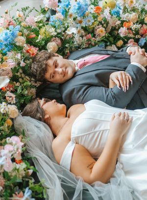 Bride and groom lying among colorful flowers, holding hands in a romantic wedding portrait.
