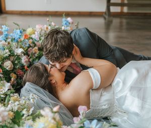 Bride and groom sharing a romantic kiss while lying among colorful floral arrangements on their wedding day.