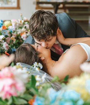 Two people sharing a romantic kiss surrounded by colorful flowers in a cozy indoor setting.