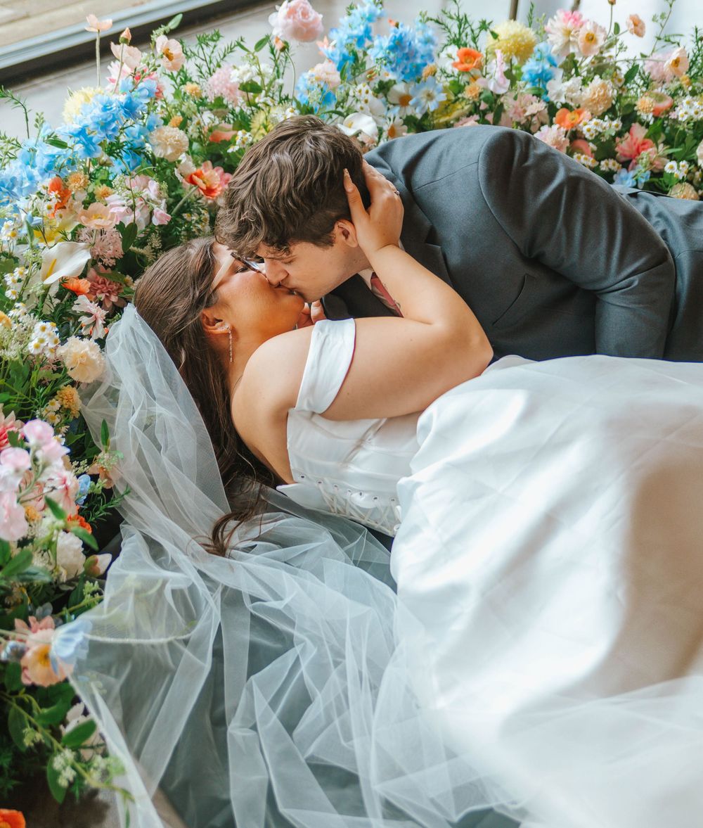 A bride and groom share a romantic kiss surrounded by colorful floral arrangements on their wedding day.
