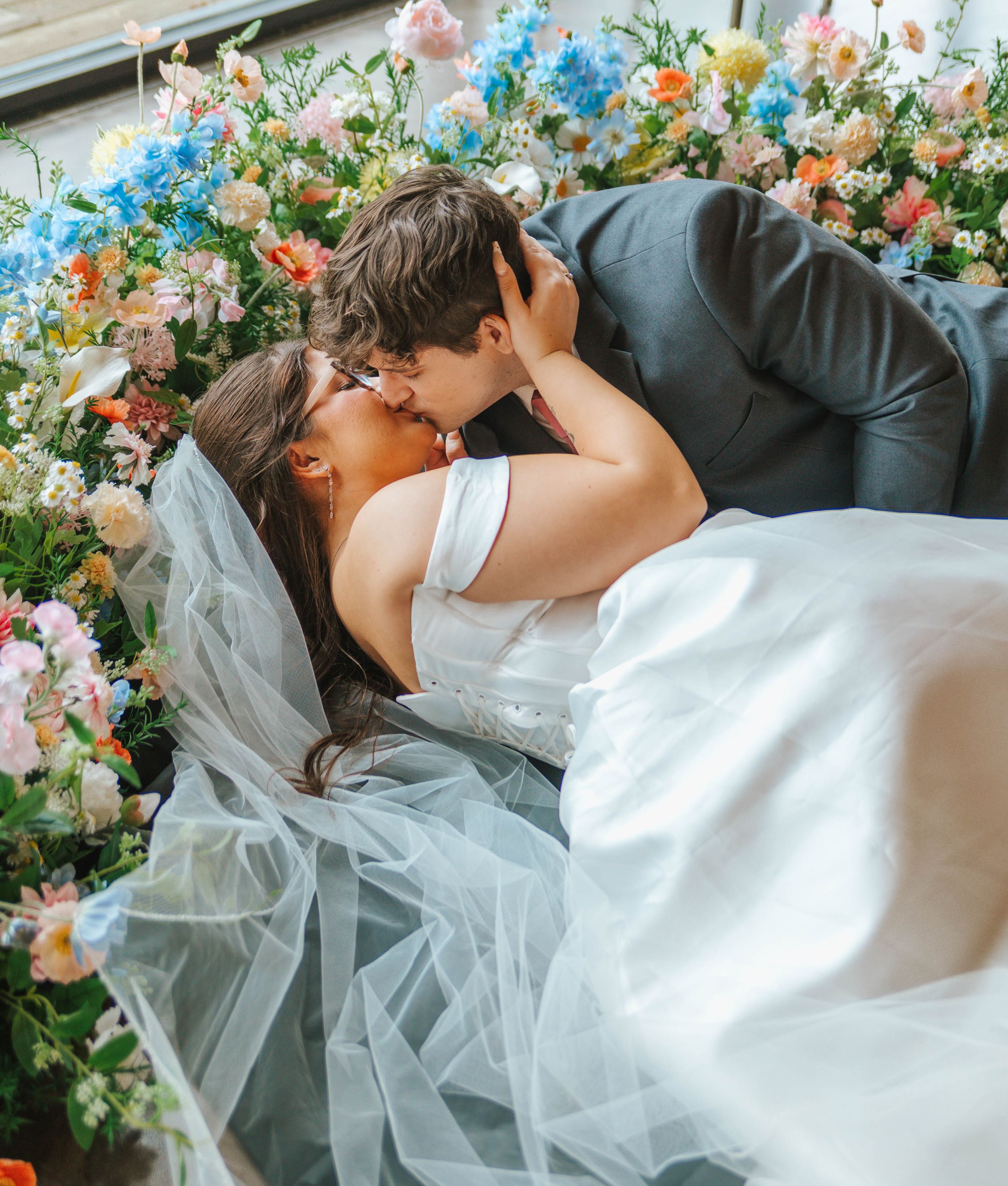 A bride and groom share a romantic kiss surrounded by colorful floral arrangements on their wedding day.