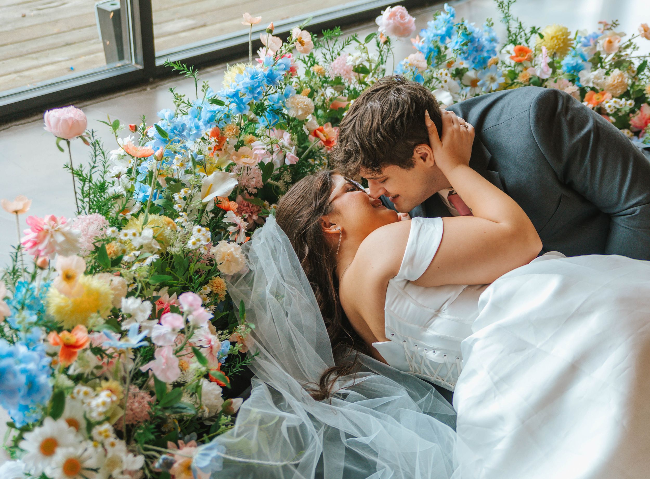 Bride and groom lying among colorful wildflowers, sharing a romantic moment in a dreamy wedding photo.