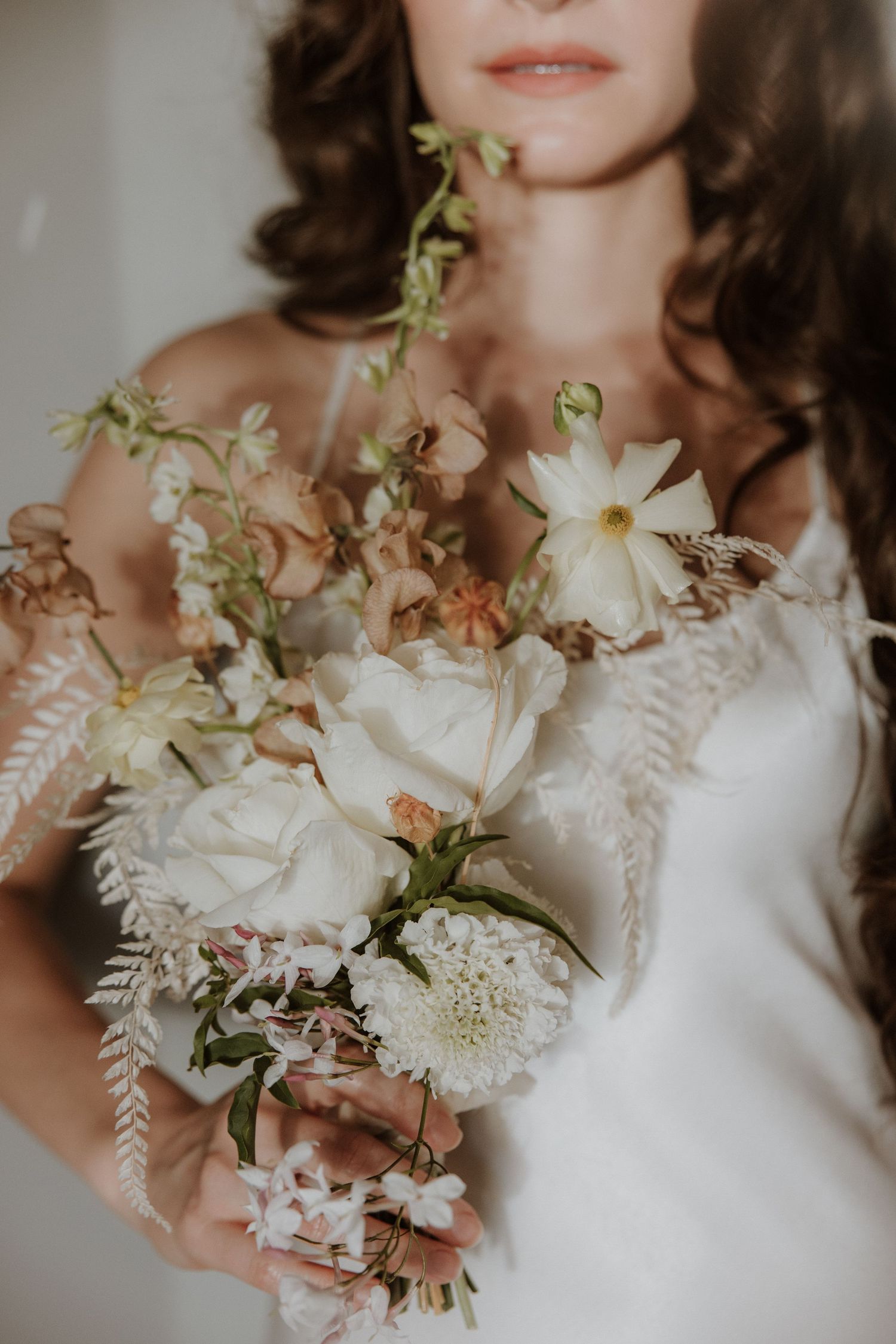 Bride in white lace gown holding a romantic bouquet of white roses, cosmos, and blush blooms with trailing greenery.