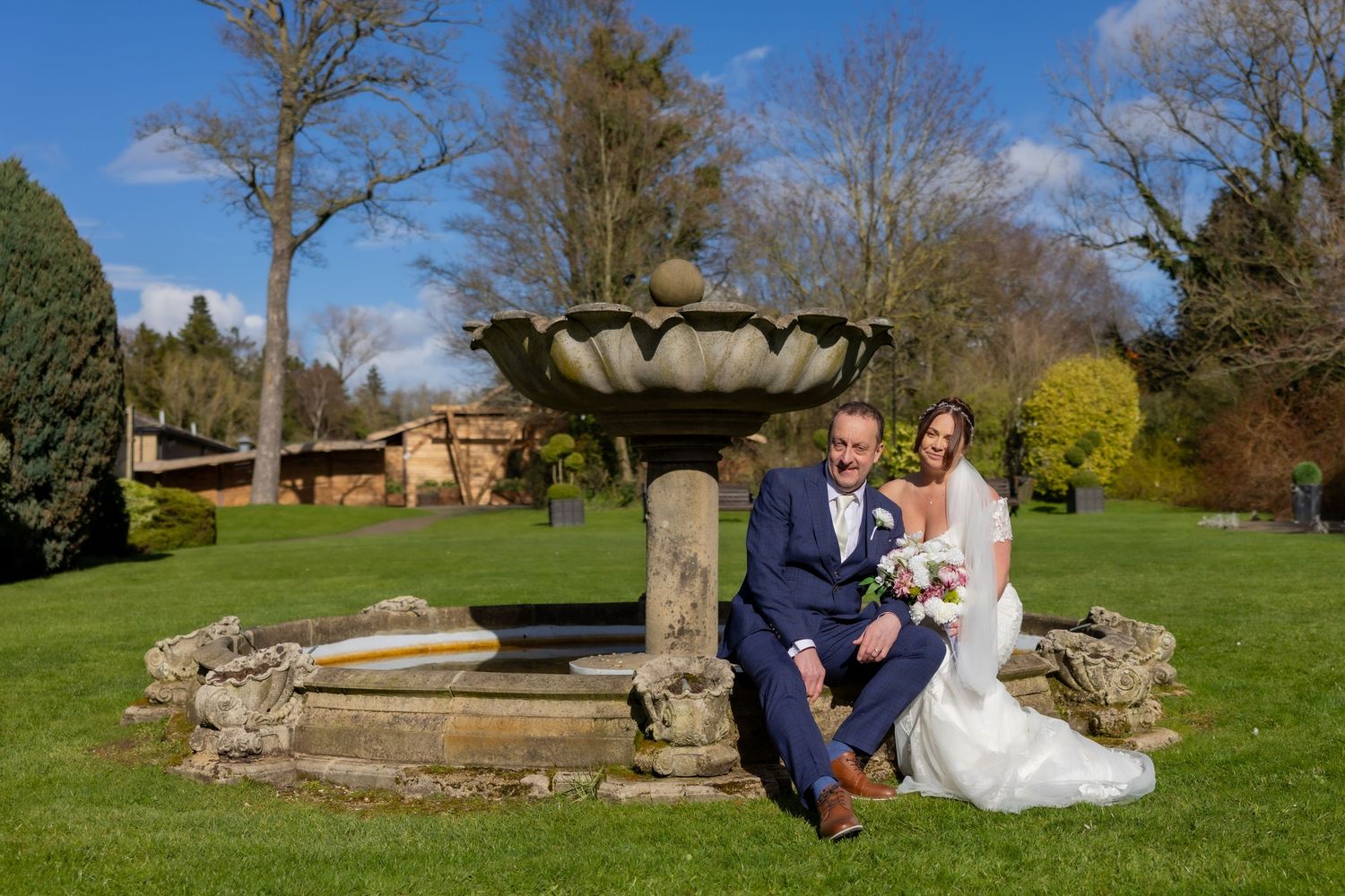 Bride and groom posing by a stone fountain in a lush garden on their wedding day. Casual Captures Wedding at Ramside Hall Hotel.